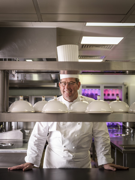 John Williams, Executive Chef at The Ritz London, stands proudly in the restaurant’s kitchen, dressed in full chef whites and a traditional toque. He smiles behind a row of cloche-covered dishes, ready for service.